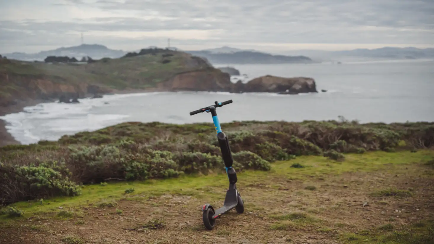 An electric scooter resting peacefully on a hilltop with cliffs and the ocean in the background.